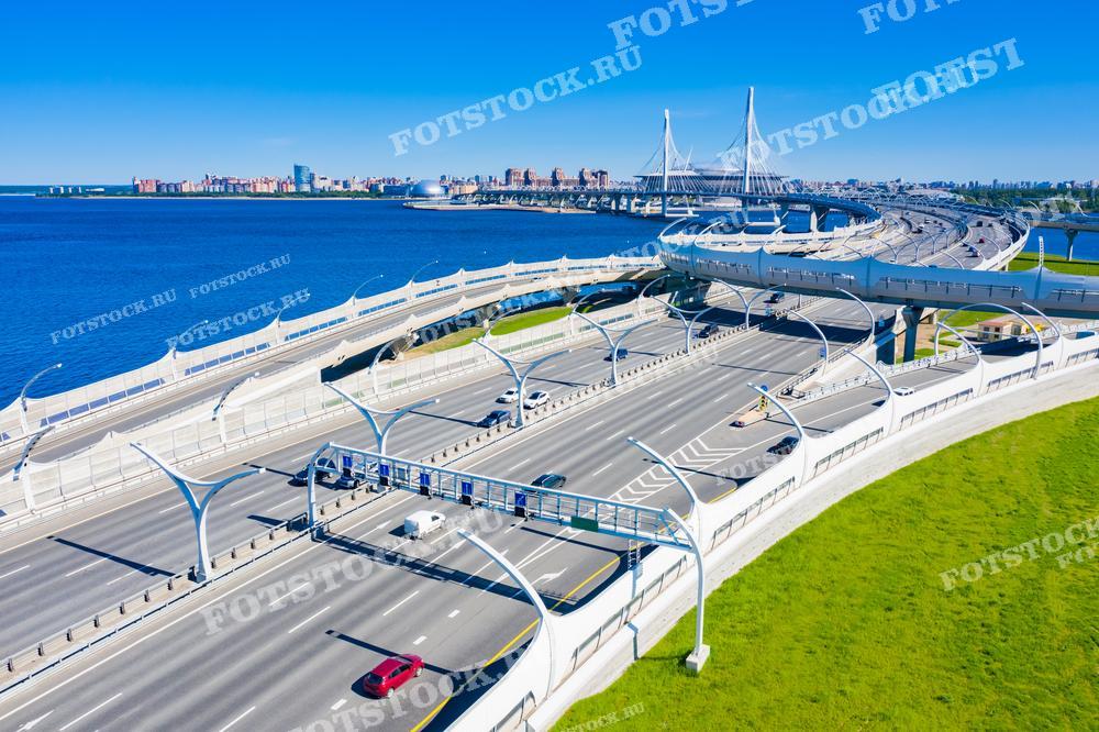 Это фотография современного моста в Санкт-Петербурге, сделанная с высоты птичьего полёта. На заднем плане видны жилые массивы и акватория Финского залива. Чистые линии дорог, яркость неба и белизна конструкций создают ощущение порядка и развитой инфраструктуры. Такое изображение подойдёт для иллюстрации статей о градостроительстве, развитии транспортной системы или как элемент графического дизайна в проектах, связанных с инженерией и строительством.